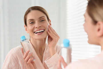 Beautiful woman removing makeup with cotton pad near mirror indoors