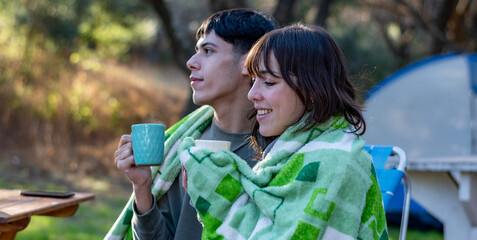 Two funny and cold friends drinking tea covered with a blanket. Campfire at the campsite