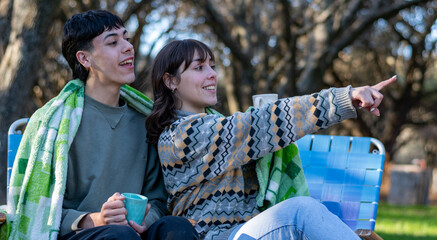 Two funny and cold friends drinking tea covered with a blanket. Campfire at the campsite