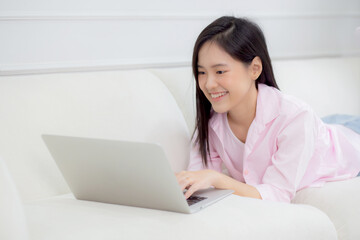 Young asian businesswoman work from home with laptop computer online to internet on sofa in living room, woman is freelance using notebook lying down on couch with comfort and relax, business concept.