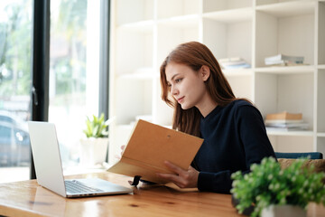 A beautiful caucasian female student is studying in college remotely. She is sitting at a table at home with a laptop and a notepad and concentrated is watching a video conference lesson
