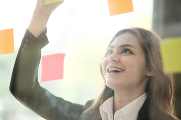 Beautiful female employee write down on colorful sticky notes manage list, concentrated biracial woman work on startup brainstorm collaborate plan on stickers on glass wall.