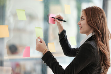 Beautiful female employee write down on colorful sticky notes manage list, concentrated biracial...