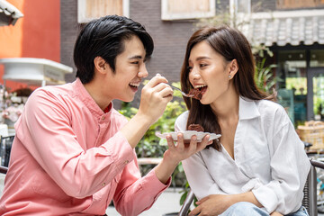 Young asian man feeding happy girlfriend with tasty chocolate cake sitting at table with coffee at date in restaurant