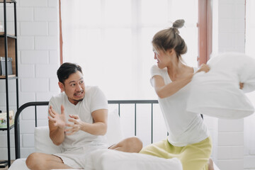 Fototapeta premium Funny asian couple angry wife hit husband with pillow in white bedroom.