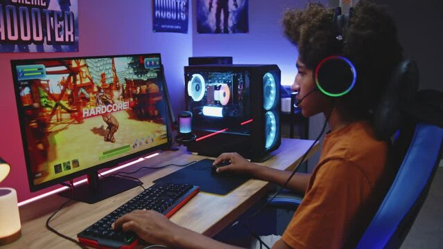 African American gamer plays in third person shooter on PC at home. Computer monitor with displayed online video game live stream or cybersport tournament. Teenager spending leisure time. Dolly shot.