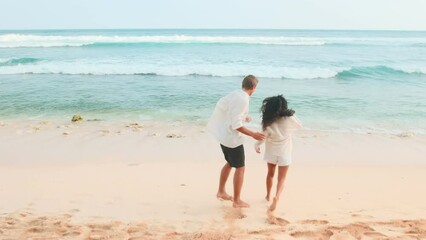 Young cheerful multiethnic friends Caucasian man and Indian woman run together along sand towards sea rejoicing at long-awaited trip to ocean and opportunity to swim located on sandy beach.