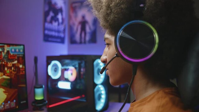 African American gamer talks with teammates and plays in third person shooter on PC at home. Online video game live stream or esports tournament. Teenager spending leisure time. Close up. Dolly shot.