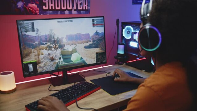 African American young gamer in headphones plays online tank battling 3D game on powerful personal computer in neon room. Video game live streaming or professional eSports tournament. Gaming at home.