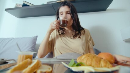 Young beautiful woman taking tray with food, having breakfast in bed in the morning, drinking coffee and enjoying it. POV zoom shot - Powered by Adobe