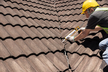 Worker man using silicone sealant adhesive  to fix crack of the old tile roof. - 621442826
