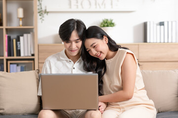 Asian young couple sit on couch or sofa looking at Computer Laptop smile and laughing together. Cheerful lover surfing internet and social media spending time in holiday at cozy home. Valentine Day