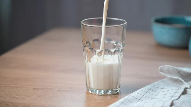 Pouring milk in a glass on wooden kitchen table. Glass with kefir or milk beverage on the table. Drinking fresh lactose-free yogurt.