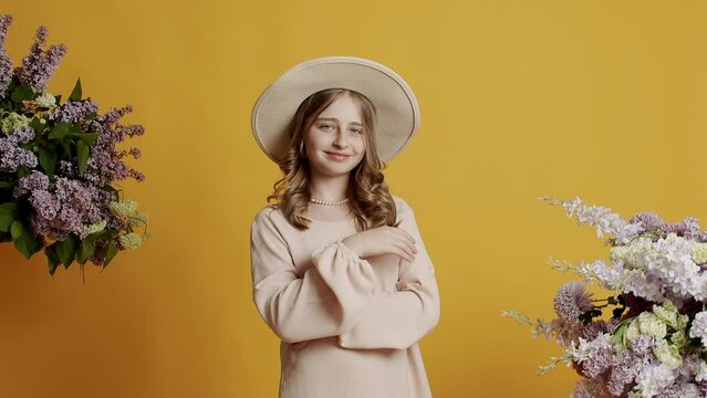 Beauty blonde teenager girl 13 years in a beige dress and hat is happy and smiling and looking at camera on a yellow background with natural flowers in the studio. Beautiful happy teenage girl smiling