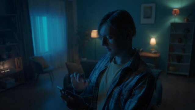 A young man in a dark living room in the evening uses a smart home control application on a smartphone. A man using a smartphone turns on the light in the living room. Smart home concept.