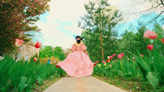 Fantasy girl princess runs in spring garden flowers tulips tree green grass alley. Woman queen, old style long pink dress with train skirt fabric fly in wind slow motion. fairy lady back rear view 4k