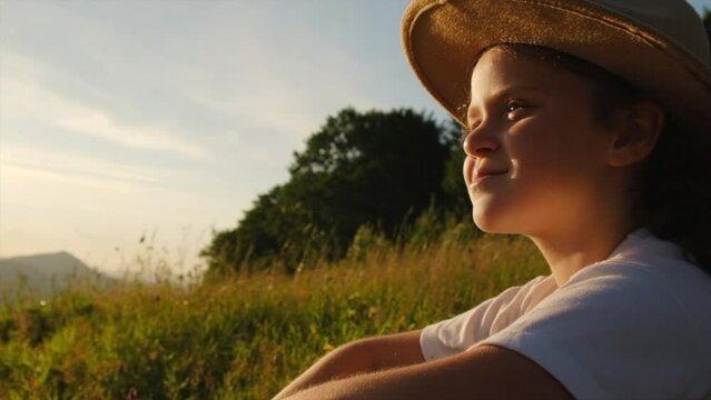 Happy teen girl in hat sitting on green grass outdoor, enjoying beautiful summer nature and mountains during warm sunset or sunrise. People emotion, travel, adventure, childhood and wanderlust concept