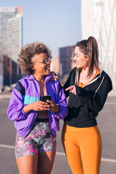 vertical photo of two sportswomen using a mobile phone before running on the city street, concept of friendship and sportive lifestyle