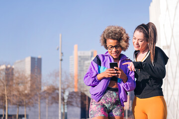 two sportswomen using a mobile phone before running standing on the city street, concept of friendship and sportive lifestyle
