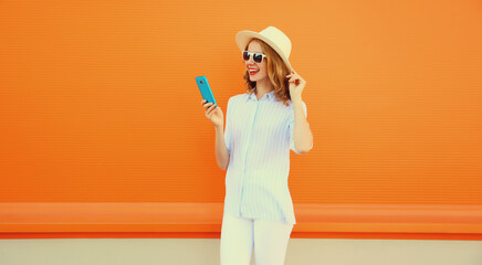Happy smiling young woman 20s with smartphone wearing summer straw hat on orange background