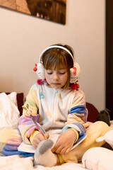 Fototapeta premium little girl in pajamas and headphones sits on the bed, draws with a pen in a notebook, vertical format