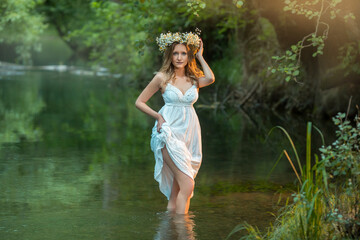 Fototapeta na wymiar a young beautiful girl in a white dress is standing in the water in the forest, with a wreath of daisies on her head.