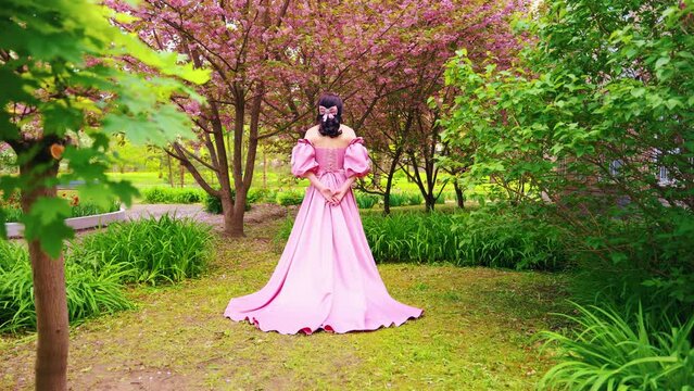 Fantasy girl princess standing in garden flowers sakura tree spring garden green grass. Sexy joy mystery woman queen back rear view long pink dress puffed sleeves satin bow in hair vintage old style