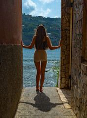 a girl stands in a narrow passage between two houses against the backdrop of the sea and mountains