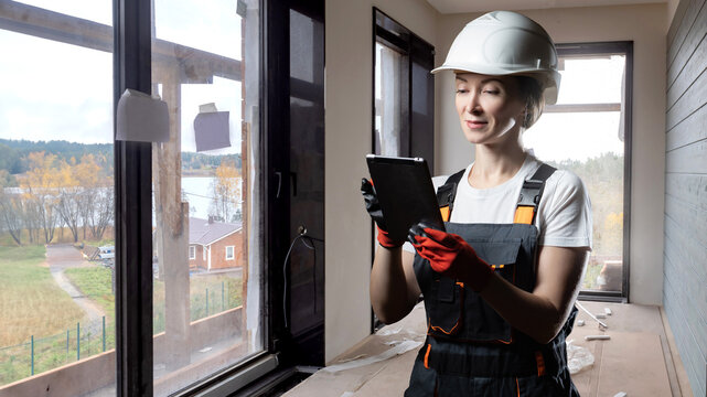 Woman builder. Girl in unfinished building. Builder with tablet in hands. Construction of building with panoramic windows. Woman in hardhat smiles. Construction worker in renovated building