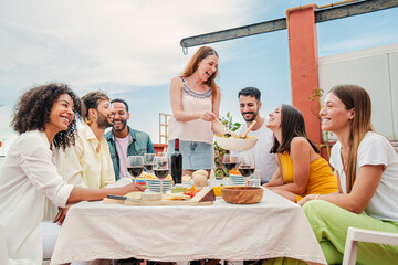 Caucasian joyful woman gathered with her friends, serving a lunch sitting at table talking and drinking wine ,enjoying a great time together. Group of young adult people celebrating at home rooftop