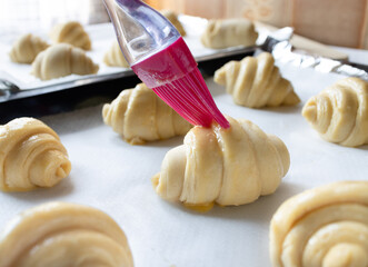 A fluffy leavened uncooked of traditional French croissants dough in a baking tray while hand holding brush doing an egg wash. With several doughs in background