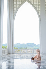 The image of an Asian Muslim woman in the Islamic religion in hijab in white color. reading the Quran, Koran Staying in a beautiful mosque, Arabic word translation: The Holy Al Quran.
