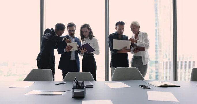 Business colleagues working in teams, using one digital gadget per group, talking, cooperating on project task, standing in modern office conference room at large window glass wall