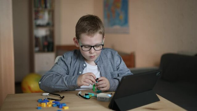 Child in room independently assembles toy from parts constructor using tablet. Boy presses on screen of gadget, shakes head negatively due to incorrect assembly of model, real time.