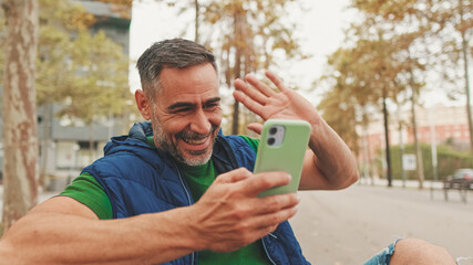Mature man wearing casual clothes, sitting on bench in the park, talking on video call from smartphone