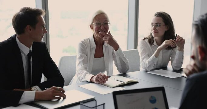 Happy confident mature older business woman talking to younger colleagues on corporate meeting, speaking, smiling, laughing, sitting ay conference table. Company teacher training interns, new workers