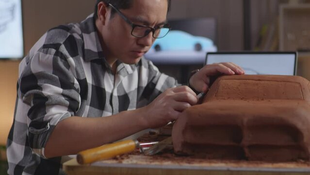 Close Up Of Asian Man Automotive Designer Looking At A Laptop And Using Rake Or Wire To Smooth Out The Surface And Create Details In The Sculpture Of Car Clay In The Studio
