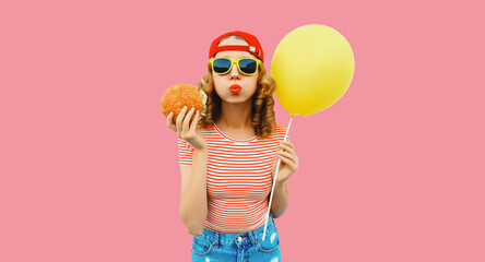 Portrait of cheerful young woman with burger fast food and yellow balloon on pink background