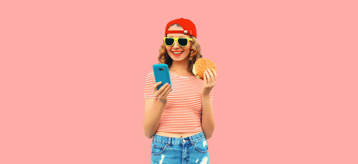 Portrait of stylish smiling young woman with phone and burger fast food on pink background