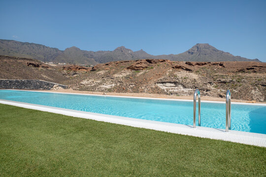 Luxurious long swimming pool overlooking the arid volcanic mountains, surrounded by a perfectly trimmed lawn, tranquil atmosphere of a high-end residential property suited for a lavishing lifestyle