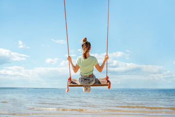 A teenage girl in jeans rides on a swing on the seashore on the beach