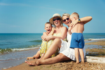 Laughing and hugging family with children on the seashore on a sunny day. Grandmother in a yellow sundress and hat, mother in a white dress and boys sons. Active lifestyle, tourism and travel.