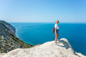 A young woman in denim shorts and a white T-shirt stands on a cliff against the backdrop of the sea and blue sky on a sunny day. Active recreation, travel and tourism.