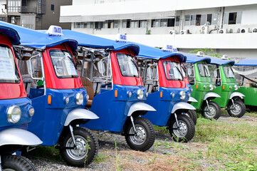 BANGKOK, THAILAND - JULY 10, 2023: Several Electric tuk tuks taxi stopped in the meadow with buildings background. Concept to save fuel, maintain and reduce air pollution protect the environment.