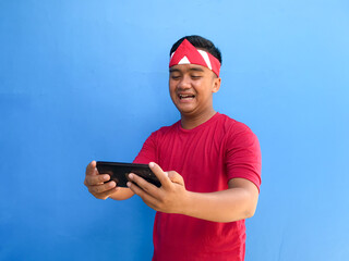 Beautiful Asian man giving salute celebrate Indonesian independence day on August 17 using red shirt isolated over blue background. Playing a mobile game and holding a smartphone. Semarak Kemerdekaan 