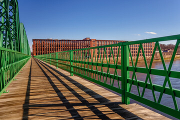 Brightly painted green bridge over river with 19th century brick mill building in background.