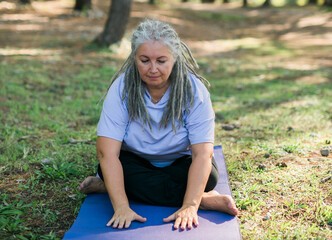 Naklejka na ściany i meble Senior woman with dreadlocks in stretching position on nature at morning. Elderly woman doing yoga on park - wellbeing and wellness tai chi