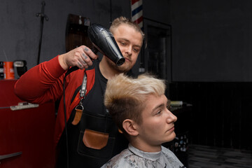 A man, a professional hairdresser, Caucasian, stylish dries the hair of a client, a guy with a hairdryer in the process of cutting at work in the salon