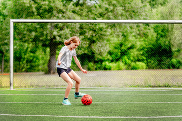 Pretty child girl running with football ball on grass during game. Cute female kid at socker field...
