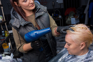 A girl is a professional barbershop worker drying her hair with a blue hair dryer for an adult client at work